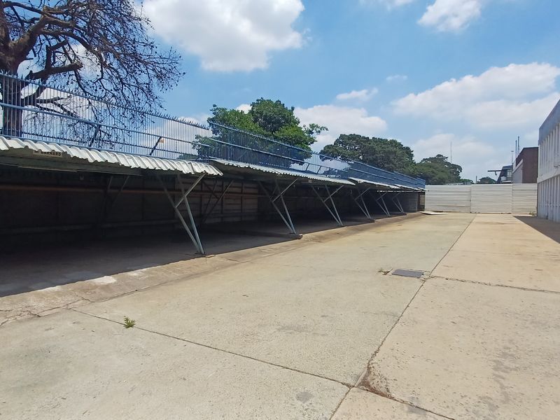 Industrial outdoor area featuring a long covered parking or storage structure with metal roofing and concrete paving adjacent to a multi-story building.