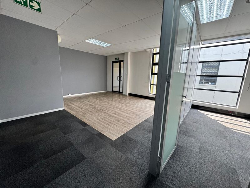 A bright, vacant office space featuring a mix of carpet and laminate flooring, glass partitioning, and large windows providing ample natural light.