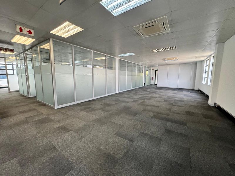 Spacious open-plan office area featuring glass partitioned offices, carpeted flooring, suspended ceilings with recessed lighting, and integrated air conditioning.