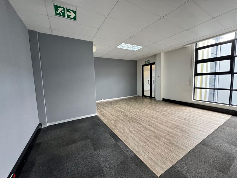 A bright, vacant office space featuring a suspended ceiling, recessed lighting, large windows, and a combination of carpet and wood-look flooring.