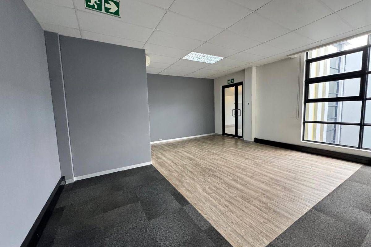 A bright, vacant office space featuring a suspended ceiling, recessed lighting, large windows, and a combination of carpet and wood-look flooring.