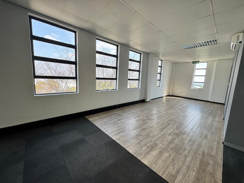 A bright, open-plan office space featuring large windows, a mix of laminate and carpet flooring, suspended ceilings, and a wall-mounted air conditioning unit.