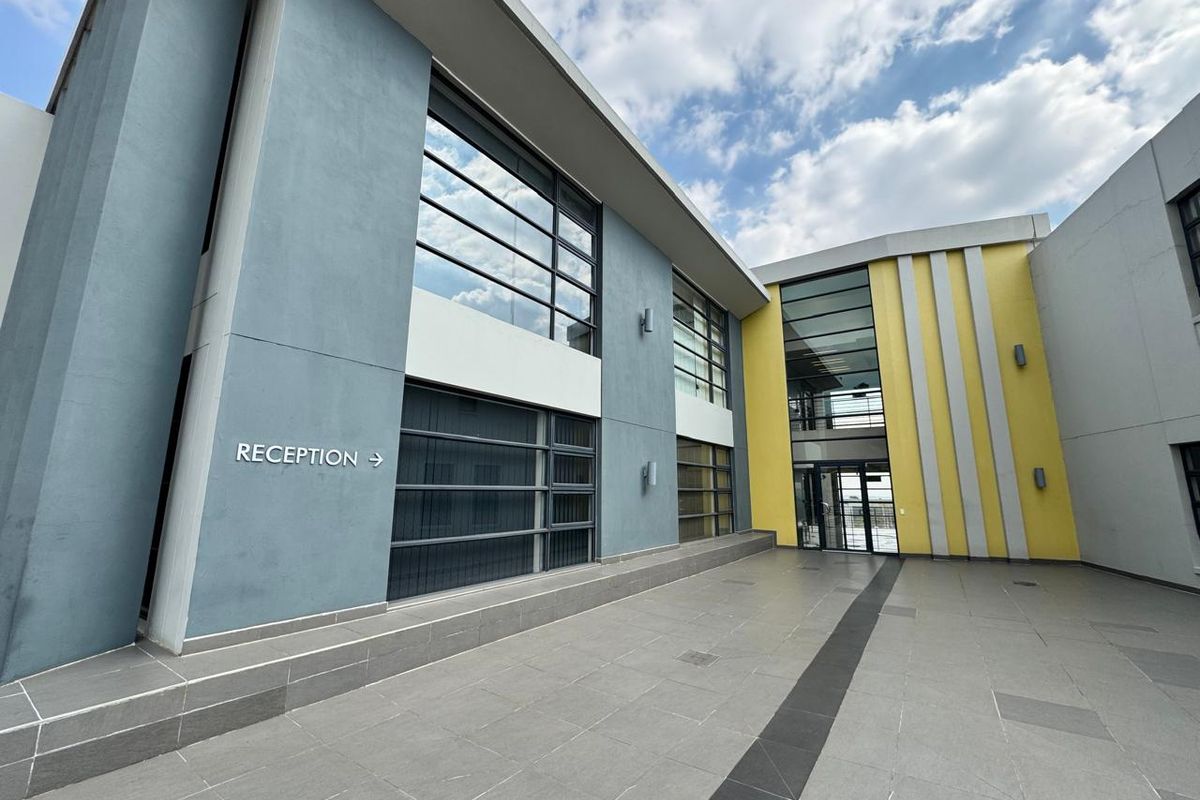 A modern office building courtyard featuring a paved walkway, large glass windows, and a clearly marked reception entrance.