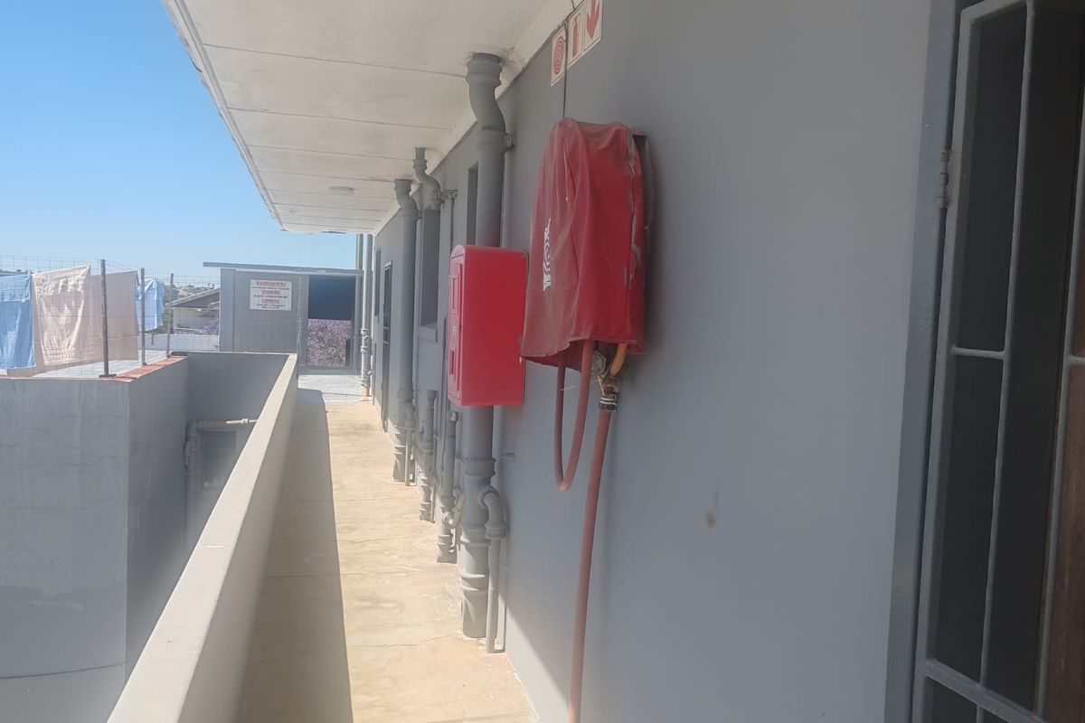 An exterior walkway featuring exposed plumbing, a fire hose reel, and a fire extinguisher cabinet, providing access to the building's rear section.