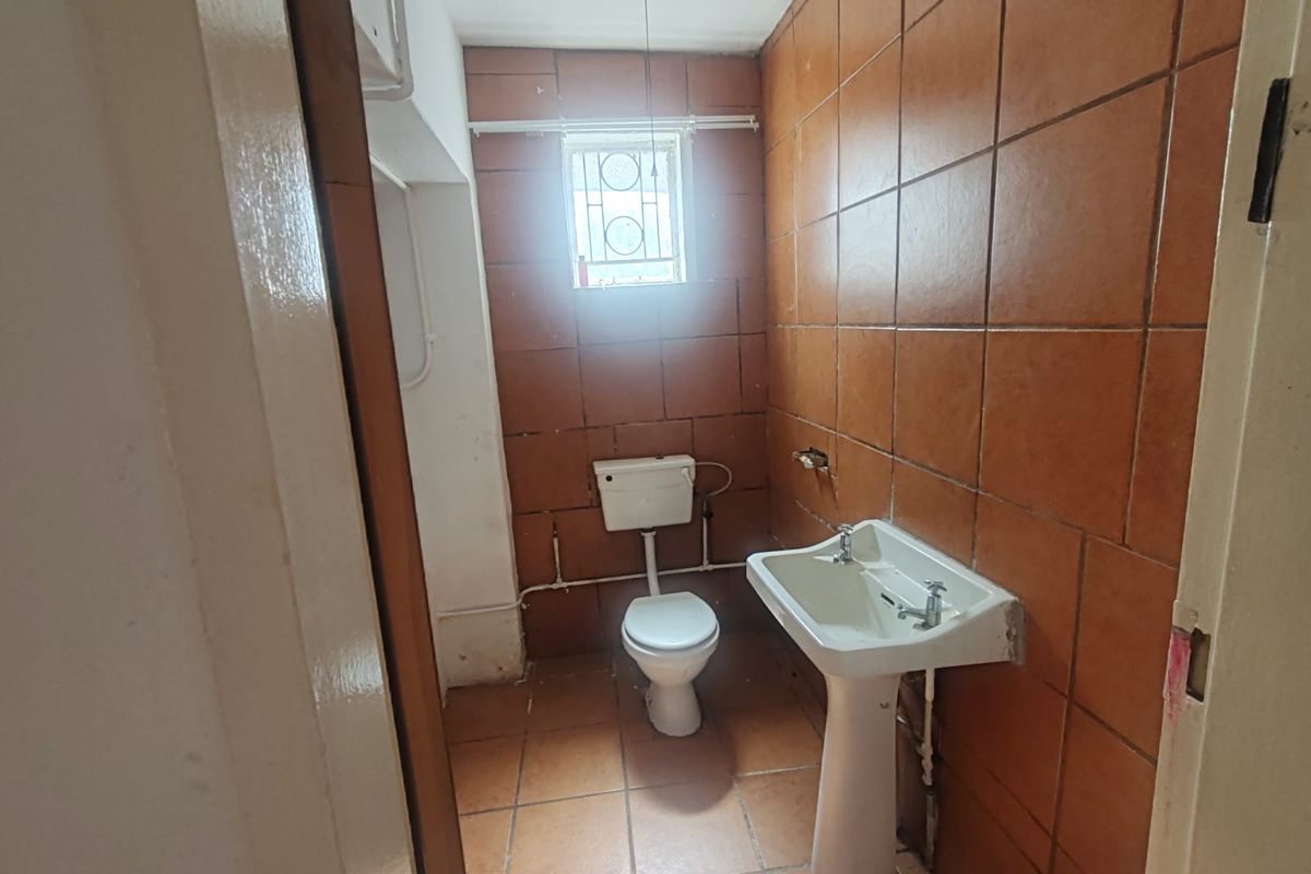 A functional commercial bathroom featuring a pedestal basin, toilet, and full-height ceramic wall tiling with a small window for ventilation.