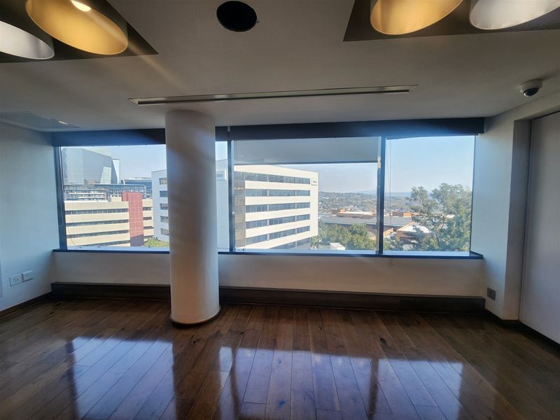 A bright, empty office space featuring polished hardwood floors, large windows with city views, and modern recessed lighting.