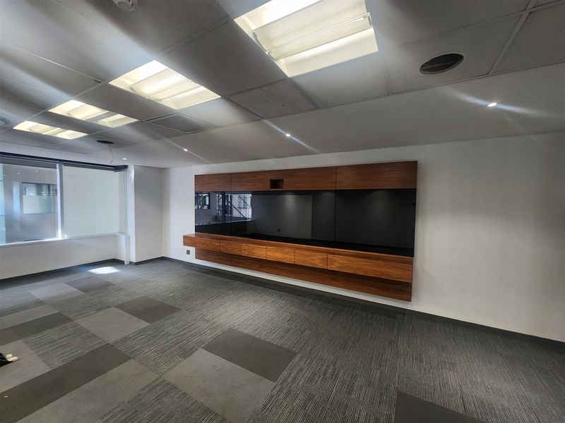 A spacious, empty office area featuring modern built-in wooden cabinetry, carpet tile flooring, and a suspended ceiling with recessed lighting.