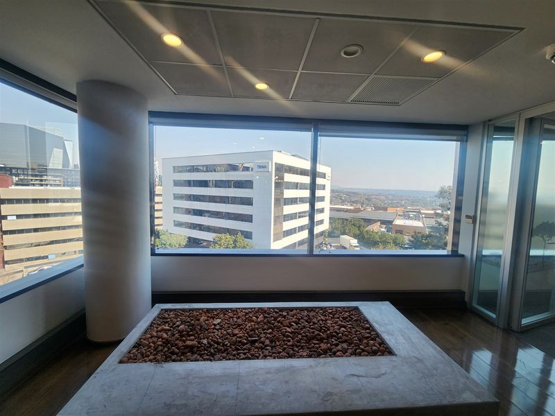 A bright, modern office space featuring expansive floor-to-ceiling windows with panoramic city views, recessed lighting, and a unique decorative stone feature.