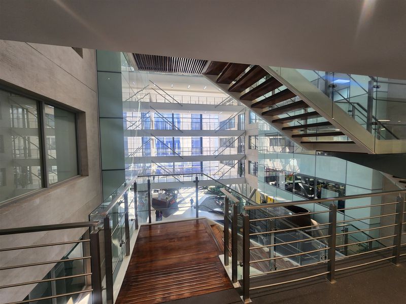A modern office atrium featuring a timber staircase with glass balustrades and extensive floor-to-ceiling glazing, creating a bright and open multi-level workspace environment.
