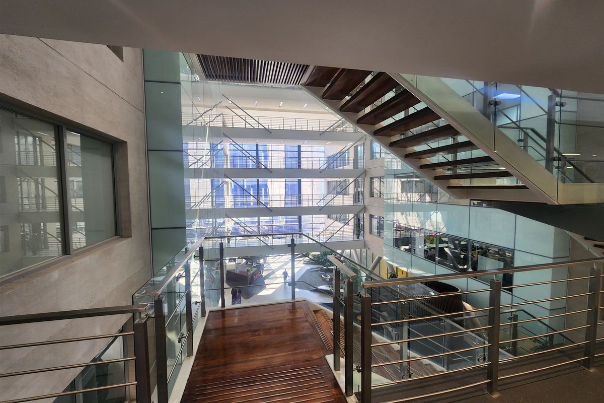 A modern office atrium featuring a timber staircase with glass balustrades and extensive floor-to-ceiling glazing, creating a bright and open multi-level workspace environment.