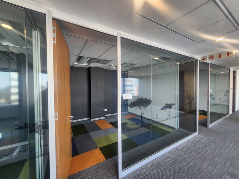 Modern office space featuring glass partition walls, suspended ceilings with recessed lighting, and colorful modular carpet tiles in a professional commercial setting.