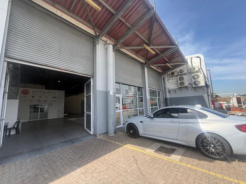 Industrial facility exterior featuring large roller shutter doors, paved driveway access, and multiple wall-mounted HVAC condenser units.