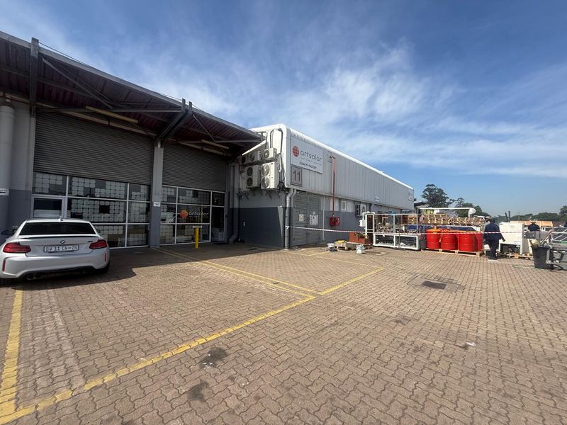 Exterior view of an industrial facility featuring a paved parking area, roller shutter doors, and wall-mounted air conditioning units.