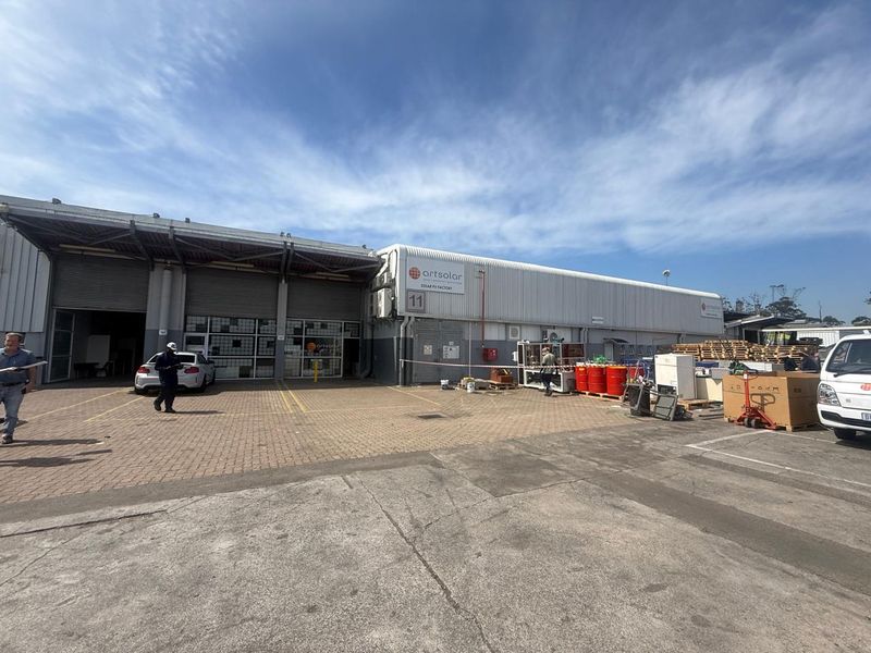 Industrial facility exterior featuring a large warehouse structure with roller shutter doors, paved loading area, and signage.