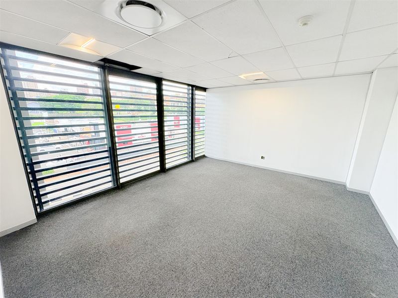 A bright, empty office space featuring large windows with security louvres, carpeted flooring, and a suspended ceiling with recessed lighting.