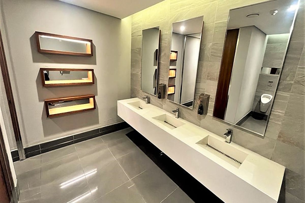 Modern commercial bathroom featuring a sleek triple vanity basin, large wall-mounted mirrors, tiled finishes, and recessed lighting.