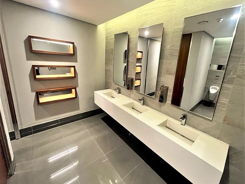 Modern commercial bathroom featuring a sleek triple vanity basin, large wall-mounted mirrors, tiled finishes, and recessed lighting.
