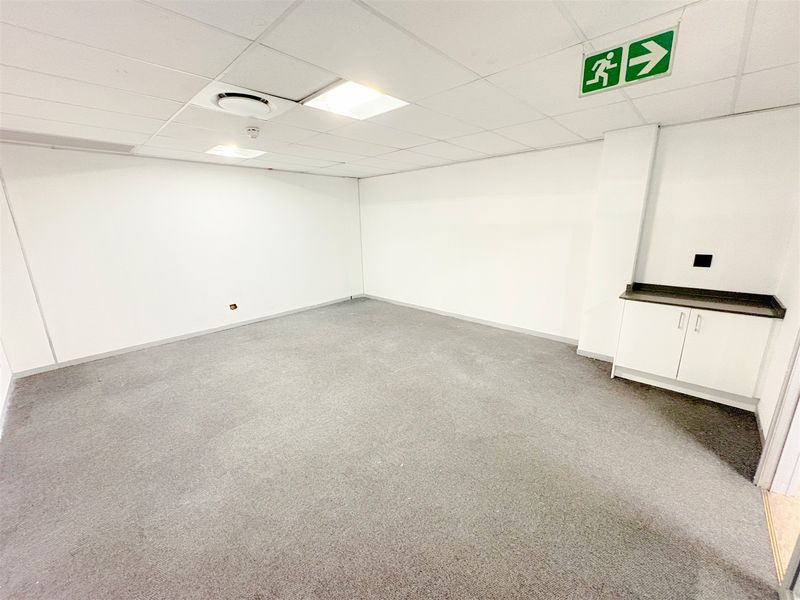 A clean, empty office space featuring neutral carpeting, a suspended ceiling with recessed lighting, and a small built-in kitchenette unit.