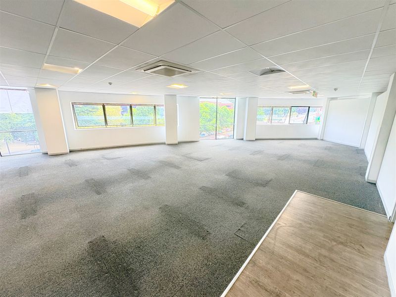 A spacious, open-plan office area featuring large windows, suspended ceilings with recessed lighting, and a combination of carpet and laminate flooring.