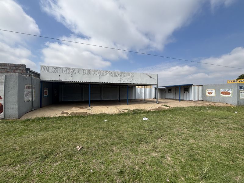 Front view of a retail butchery premises featuring a concrete forecourt, metal roller shutter doors, and corrugated metal construction.