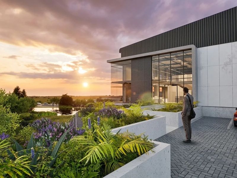 Modern industrial building exterior featuring glass facade, concrete planters, and paved driveway overlooking a landscaped garden at sunset.