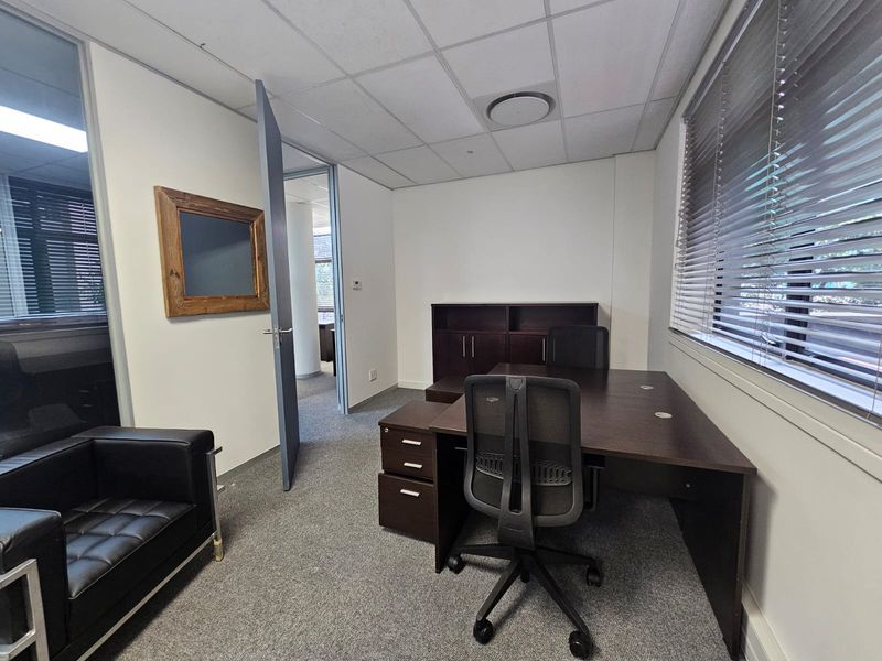 A functional office space featuring carpeted flooring, a suspended ceiling, and large windows with venetian blinds, providing a professional work environment.