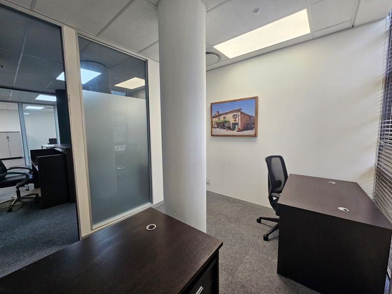 A professional office space featuring a glass partition wall, carpeted flooring, and a central structural column with recessed ceiling lighting.