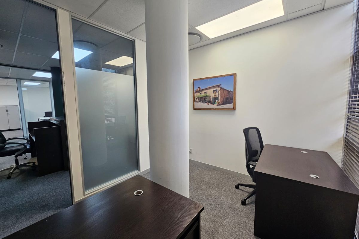 A professional office space featuring a glass partition wall, carpeted flooring, and a central structural column with recessed ceiling lighting.