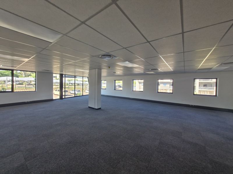 A spacious, open-plan commercial office area featuring a suspended ceiling, recessed lighting, carpeted flooring, and large windows providing ample natural light.