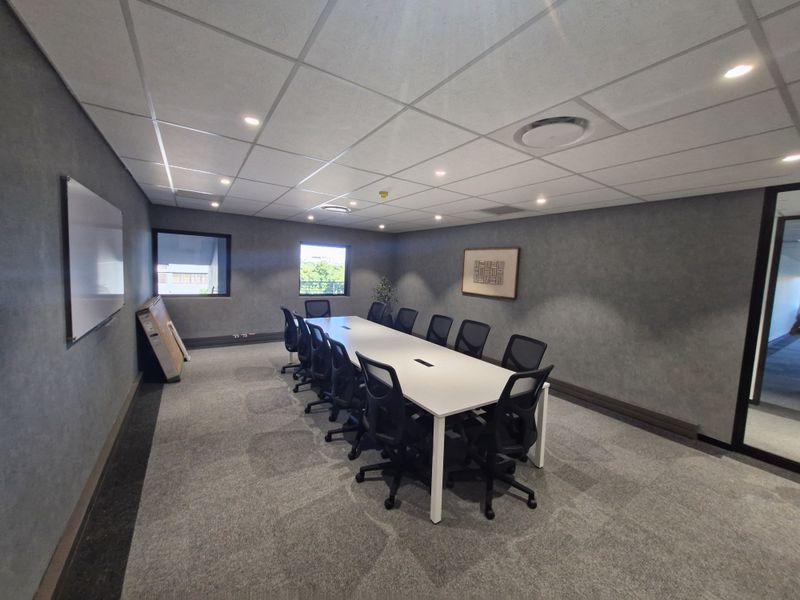 A professional boardroom or meeting room featuring a large central table, carpeted flooring, suspended ceiling with recessed lighting, and a wall-mounted whiteboard.