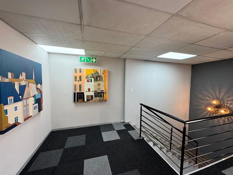A clean, modern office hallway featuring carpet tiles, a suspended ceiling with recessed lighting, and a staircase with a contemporary metal balustrade.