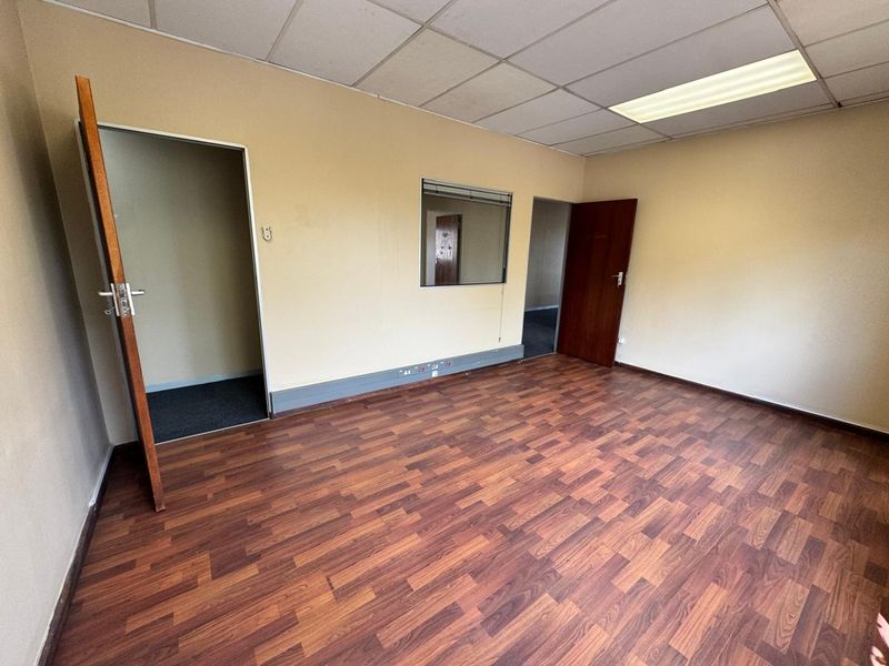 An empty office space featuring laminate flooring, a suspended ceiling with recessed lighting, and an internal window connecting to an adjacent room.