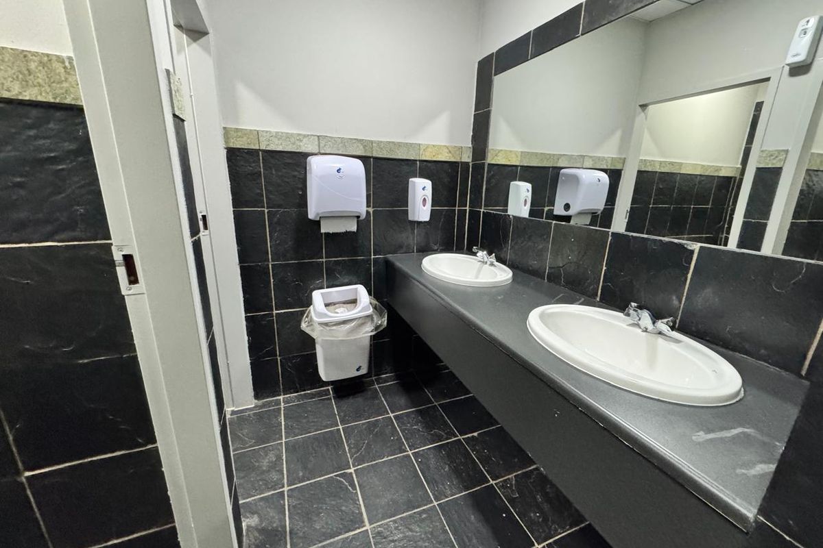 A functional office bathroom featuring dual vanity basins, a large wall mirror, and dark tiled finishes throughout.
