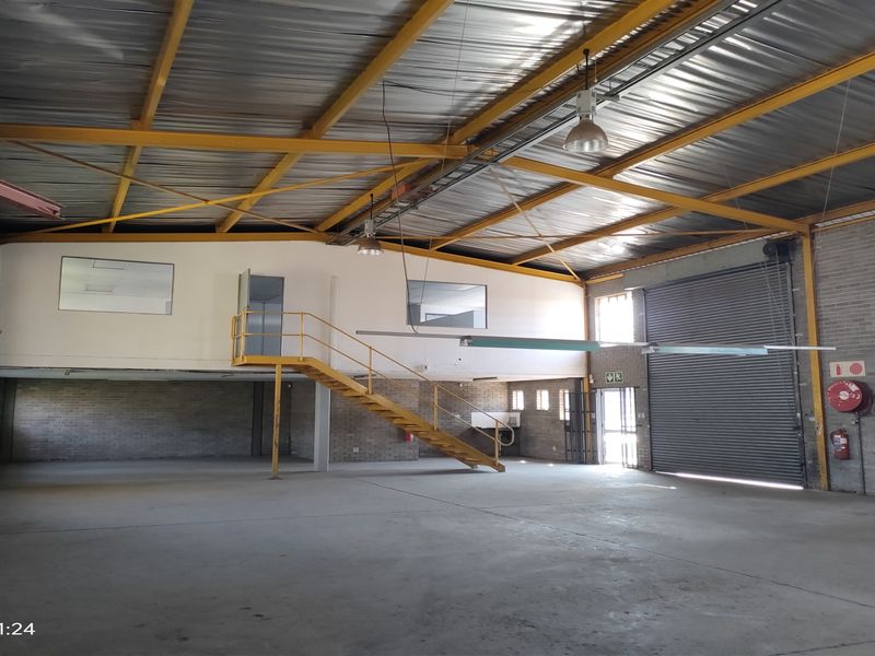 Spacious industrial warehouse featuring high ceilings with exposed steel trusses, a mezzanine level with staircase access, concrete flooring, and a large roller shutter door.