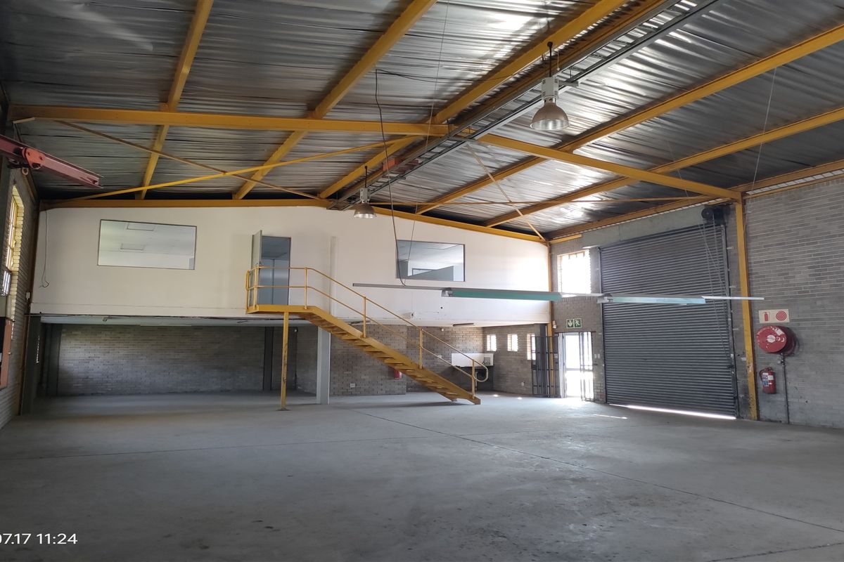 Spacious industrial warehouse featuring high ceilings with exposed steel trusses, a mezzanine level with staircase access, concrete flooring, and a large roller shutter door.