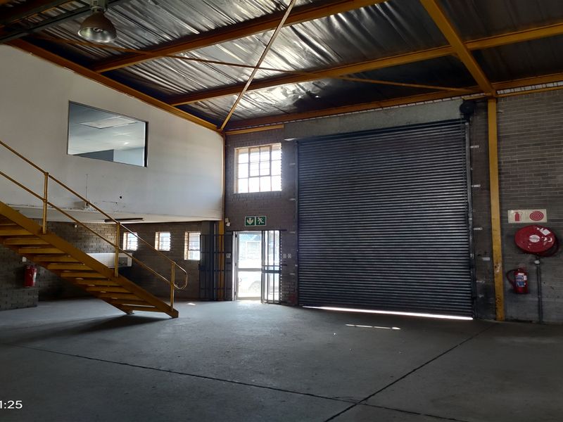 Spacious industrial warehouse unit featuring high ceilings with exposed steel trusses, a large roller shutter door, concrete flooring, and an internal steel staircase.
