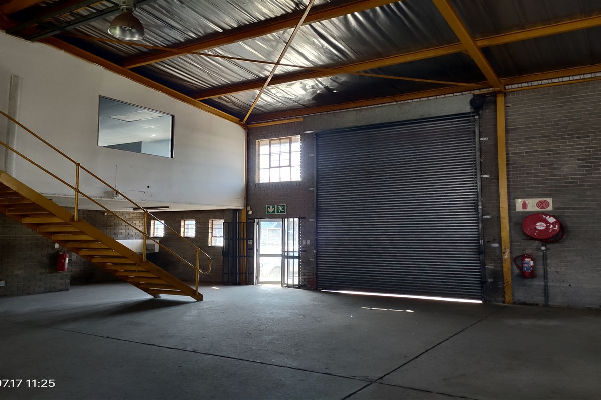 Spacious industrial warehouse unit featuring high ceilings with exposed steel trusses, a large roller shutter door, concrete flooring, and an internal steel staircase.