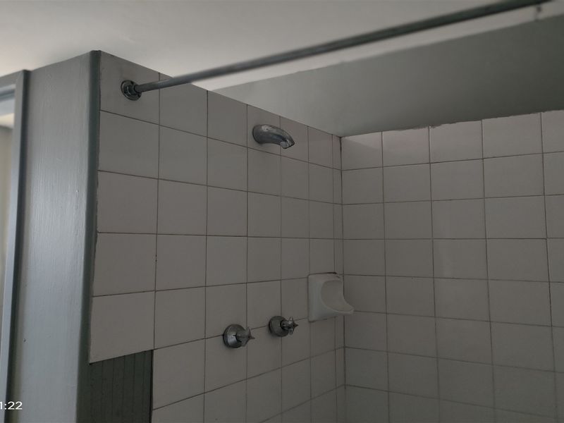 A functional shower area featuring white wall tiles, a shower head, and mixer taps, currently showing signs of wear and missing tile sections.