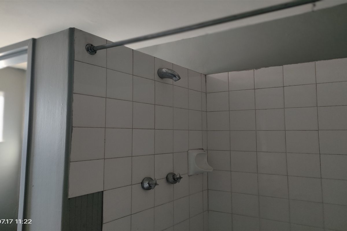 A functional shower area featuring white wall tiles, a shower head, and mixer taps, currently showing signs of wear and missing tile sections.
