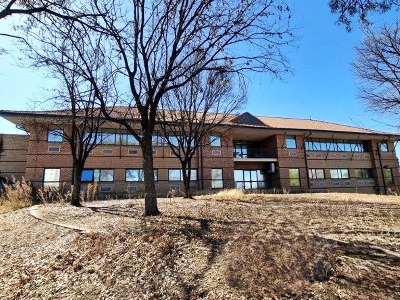 A two-story brick office building featuring a tiled roof, large windows, and a central balcony, set within a landscaped property.
