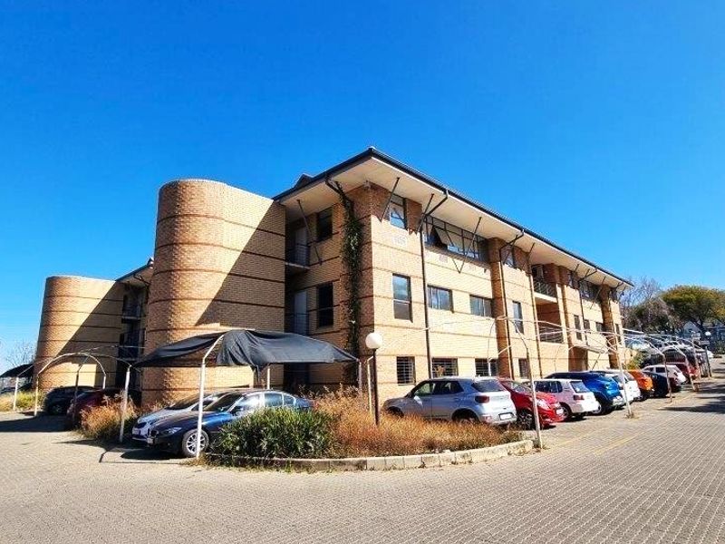 Exterior view of a multi-story brick office building featuring a paved parking area with covered bays and established landscaping.