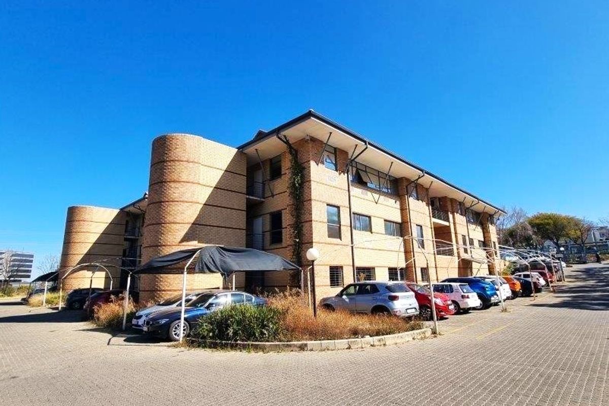 Exterior view of a multi-story brick office building featuring a paved parking area with covered bays and established landscaping.