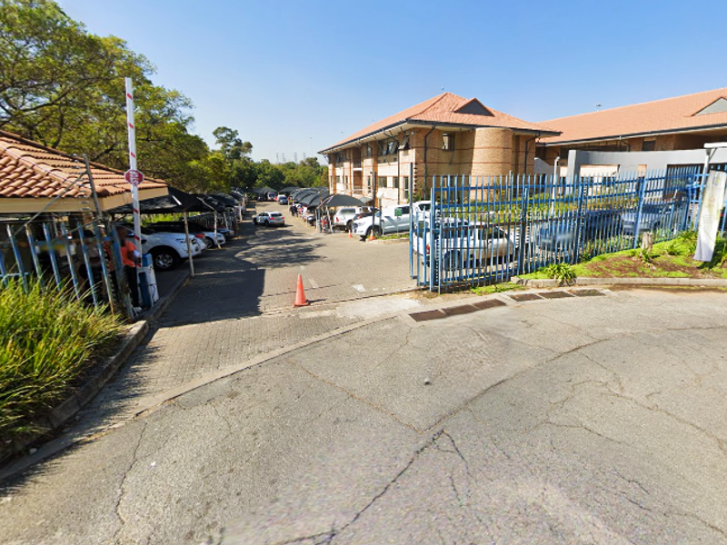 Exterior view of the office property entrance featuring a paved driveway, security fencing, access control gate, and covered parking areas.