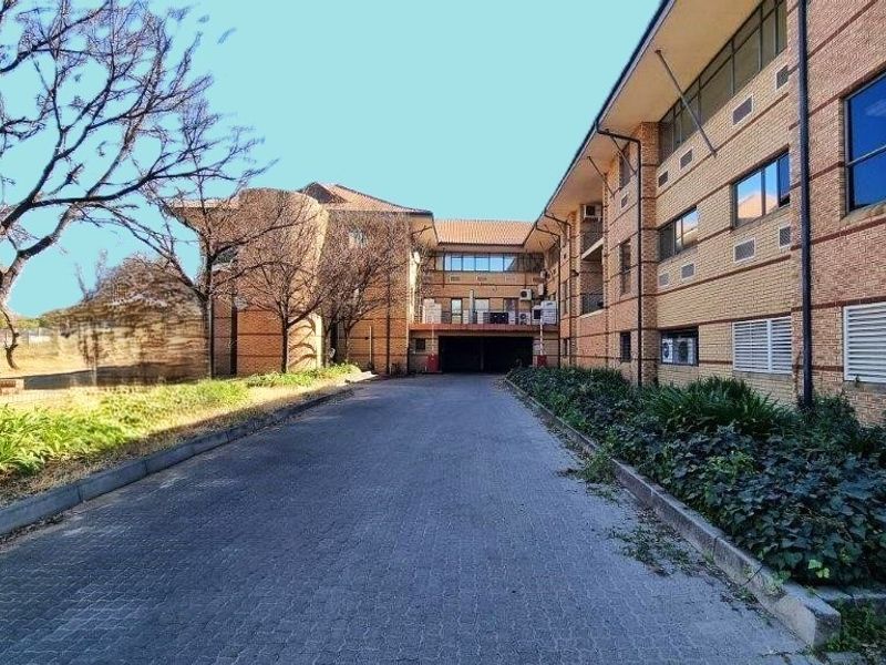 Rear view of a multi-story brick office building featuring a paved driveway, covered parking access, and established garden beds.