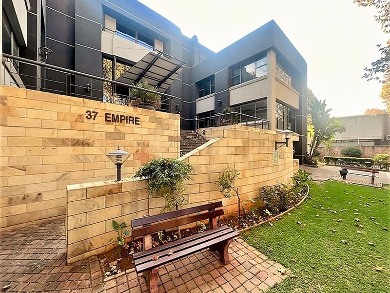 Modern office building exterior featuring a sandstone retaining wall, paved entrance walkway, and contemporary architectural facade with metal railings.