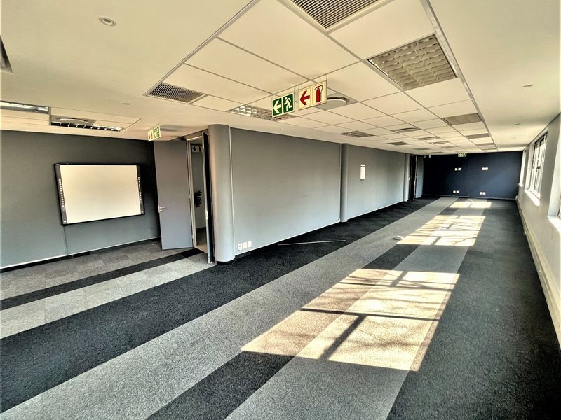A spacious, empty open-plan office area featuring a suspended ceiling, recessed lighting, air conditioning vents, and modern grey-toned carpeted flooring.