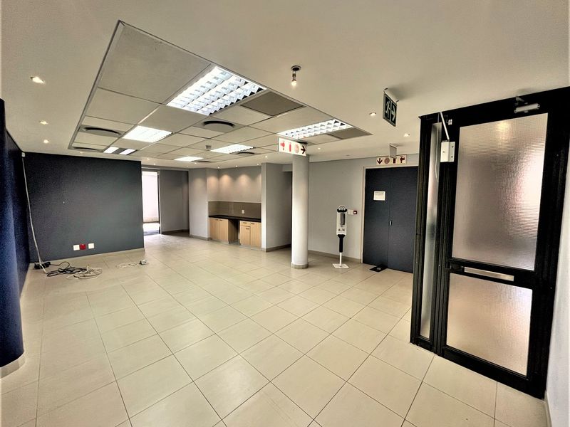 Spacious open-plan office area featuring tiled flooring, a suspended ceiling with recessed lighting, and a small built-in kitchenette unit.