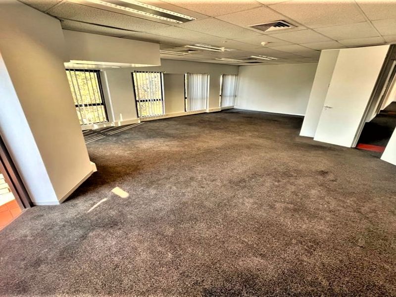 An open-plan office space featuring suspended ceilings, recessed lighting, vertical blinds, and carpeted flooring, offering a functional layout for professional use.