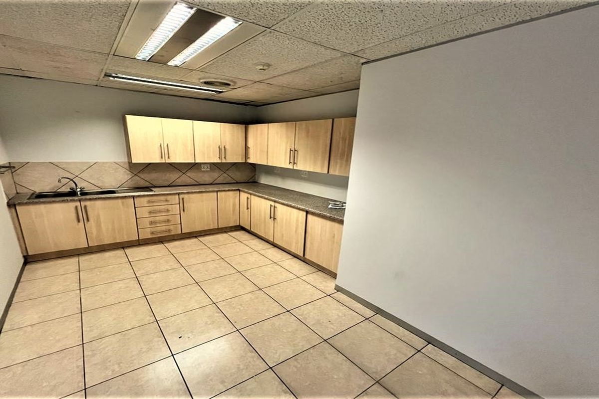 A functional office kitchenette featuring light wood-tone cabinetry, laminate countertops, a stainless steel sink, and durable tiled flooring.