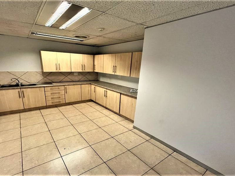 A functional office kitchenette featuring light wood-tone cabinetry, laminate countertops, a stainless steel sink, and durable tiled flooring.